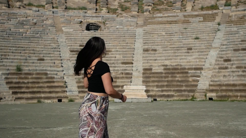 Brunette woman moves waist long lash hair. Rear view. Girl shakes long straight hair. Female model is fluttering hair. Slow motion. Ancient historical amphitheater in the background, Bodrum, Turkey. 