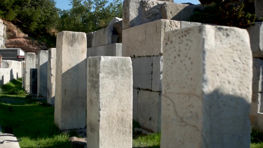 Young woman with long brunette hair walks towards a camera between antient stone blocks in the antique amphitheatre in Bodrum, Turkey. Black T-shirt, top, flourish long wide skirt pants, culottes. 