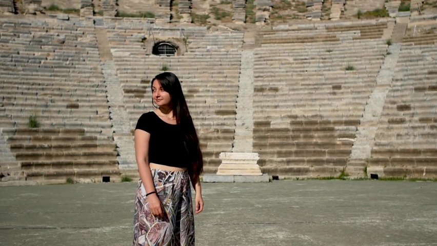 Attractive young woman looks around  with the old amphitheatre in Bodrum, Turkey. Tourist, tourism, excursion. Gladiator. Waist long brunette hair, black top, bright pans. Medium wide shot. Slowmo