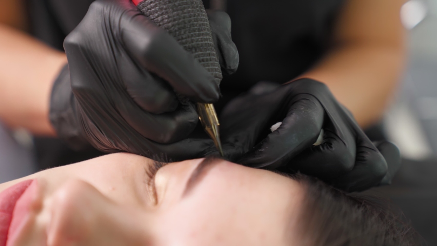 Master makes permanent eyebrow makeup procedure using special needle tattoo machine to woman in beauty salon. Microblading brows tattooing. Dark pigment is injected under skin. Cosmetology procedure Royalty-Free Stock Footage #1084171252