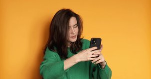 Smiling woman in sweater posing isolated on yellow background in studio. Look coquettish hold using mobile cell phone. Girl flirting while chatting on mobile phone, dreaming looks up at the camera.
