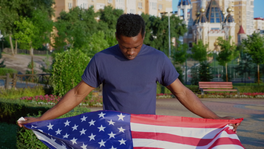 Camera zoom in patriot African American man holding an American flag and looks camera in summer. Portrait African adult male stands in park with USA flag. 4th July America independence day holidays