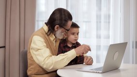 Cheerful and modern grandfather with his grandson is sitting with a laptop. An older man teaches a little boy to work on a computer, or talks over a video connection.