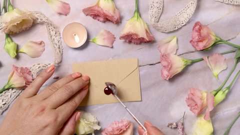 Hands pouring melted wax onto an envelope near pink flowers over a marble table close up 