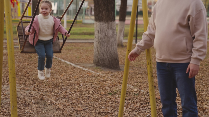 mother shakes little child on playground swing, cheerful kid flies up and down, baby laughs and smiles while playing, mother and daughter on walk in city park, happy family, childhood dream of flying Royalty-Free Stock Footage #1084180021