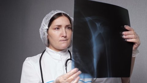 Professional lady orthopedist in uniform looks at condition of bones and joints depicted on x-ray picture standing in grey clinic office closeup.