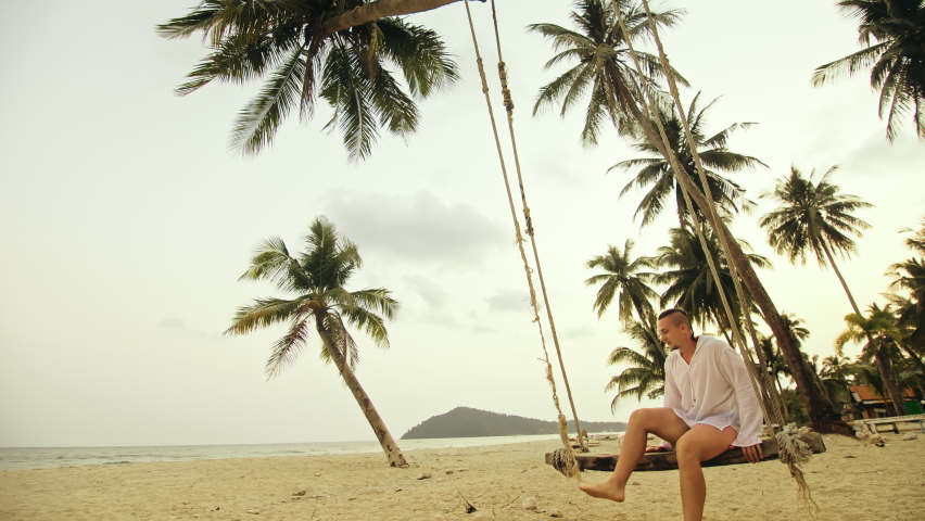 Man on the swing on tropical beach. The guy in white shirt enjoy life and has fun. Summer holiday vacation concept