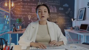 Ambitious Asian business woman is making presentation during online video call from office talking and pointing at charts sitting at desk at night