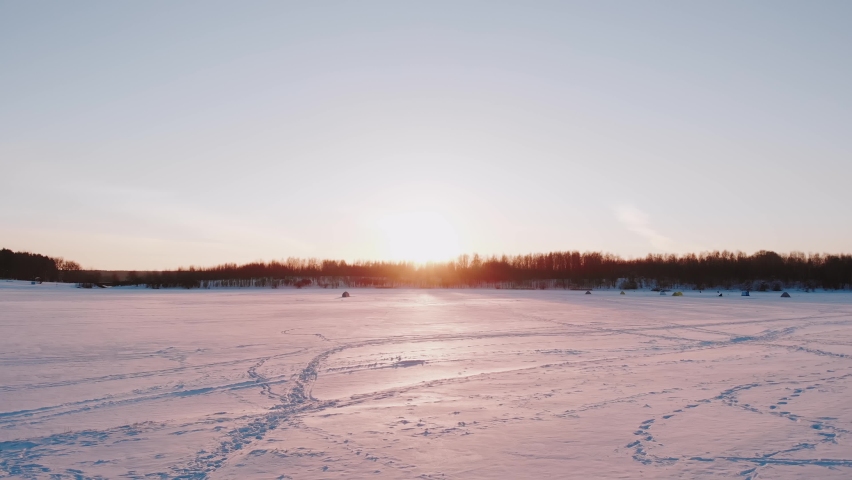 Tents of fishermen, scientists stand on frozen ice lake near forest in rays of setting sun. Surface of reservoir in footprints of people. Beautiful scenic plan of nature on frosty winter evening.