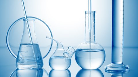 Long dropper is lowered into clear flask with water surrounded by lab glassware on pale blue background | Abstract skin care cosmetics formulation concept
