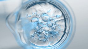 Light blue liquid pouring into beaker with water from flask creates different sized bursting bubbles and ripples on its surface on grey background | Abstract skin cleanser mixing concept - Powered by Shutterstock - Get 15% off with code: PIKWIZARD15