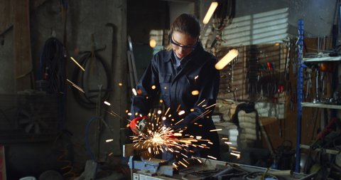 Cinematic shot of young professional female blacksmith or welder in uniform and protective goggles is using angle grinder to polish metal with sparks flying off it in her workshop.