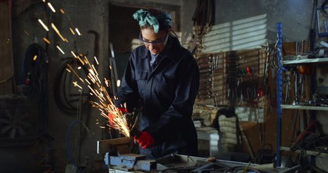 Cinematic shot of young professional female blacksmith or welder in uniform and bandana bow is using angle grinder to polish metal with sparks flying off it in her workshop.