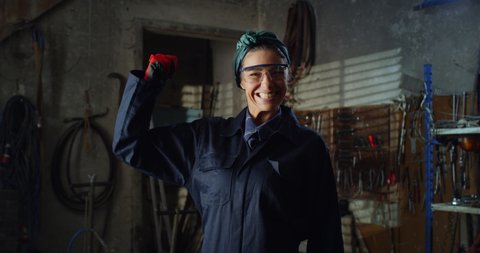 Cinematic shot of young professional female blacksmith or welder in uniform and bandana bow is using angle grinder to polish metal with sparks flying off it in her workshop.