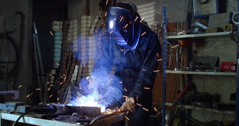 Cinematic close up shot of young professional female blacksmith or welder wearing uniform, protective helmet and gloves is working on welding metal frame with sparks flying off it in her workshop.