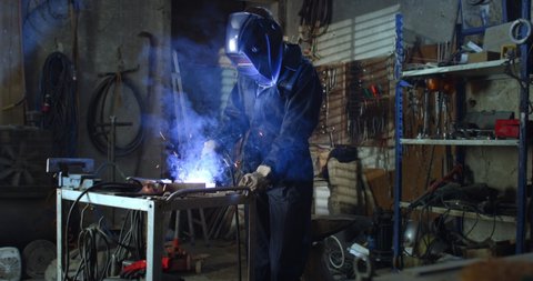 Cinematic shot of young professional female blacksmith or welder wearing uniform, protective helmet and gloves is working on welding metal frame with sparks flying off it in her workshop.