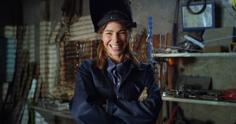 Cinematic shot of young professional female blacksmith or welder in uniform and gloves is taking off protective helmet and smiling in camera while working arc welding on metal in her workshop.
