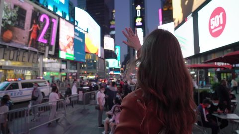 NEW YORK, USA - MAY, 18, 2021: Woman at Times square stands and looks around at night, street of Manhattan. Lots of advertising signs and glowing screens. Young girl in jacket tourist raise hand up