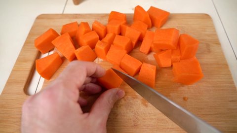 Male hands chop raw pumpkin for cooking, cut into cube pieces with knife. Cooking healthy meal from fall vegetables at rustic wooden cutting board. 