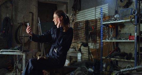 Cinematic shot of young happy smiling female mechanic in uniform is making technology video call to friends or family while having break from working in maintenance repair workshop.