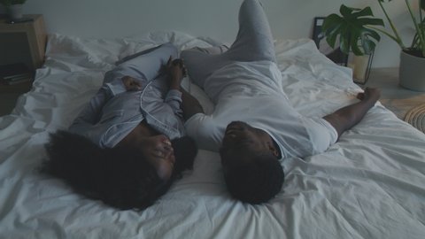 Young happy african american loving couple wearing pajamas relaxing in bed, holding hands and laughing, enjoying pastime together, top view, tracking shot, slow motion