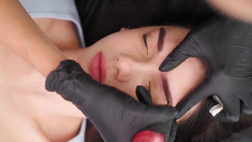 Master makes permanent eyebrow makeup procedure using special needle tattoo machine to woman in beauty salon. Microblading brows tattooing. Dark pigment injected under skin. Vertical video, top view | Shutterstock HD Video #1084212499