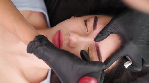 Master makes permanent eyebrow makeup procedure using special needle tattoo machine to woman in beauty salon. Microblading brows tattooing. Dark pigment injected under skin. Vertical video, top view