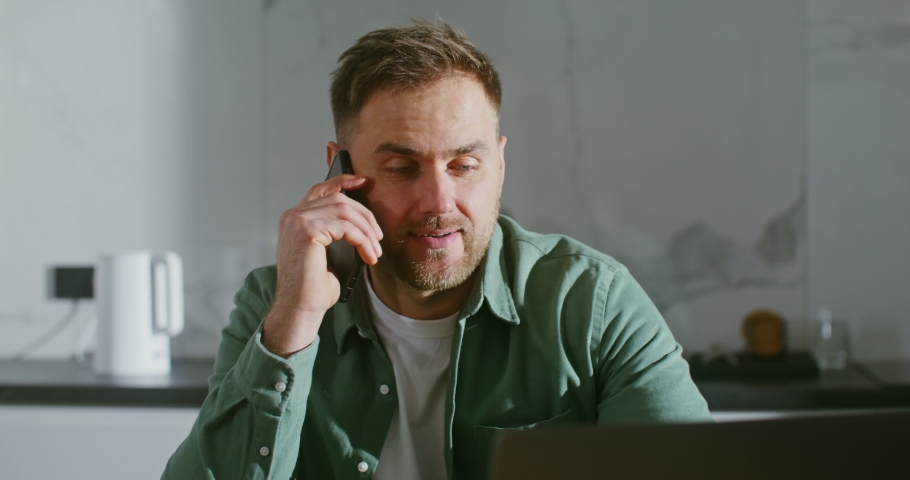 Young handsome man of European appearance with a light beard, talking on a mobile phone, looking into a laptop screen, sitting at a table in a modern stylish kitchen Royalty-Free Stock Footage #1084215910