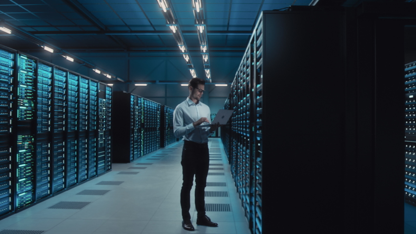 Big Data Center IT Specialist Using Laptop Computer. Server Farm Cloud Computing Facility with System Administrator Working. Software as Solution, Cyber Security. Moving Between Rack Raws Dolly Shot - Powered by Shutterstock - Get 15% off with code: PIKWIZARD15