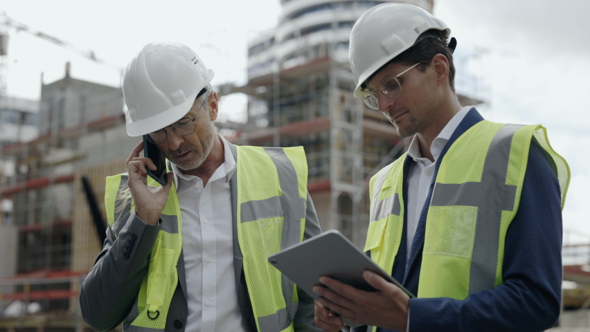 Two caucasian engineers in white safety helmets using smartphone and tablet during meeting on construction site. Concept of teamwork and modern gadgets. Royalty-Free Stock Footage #1084221679