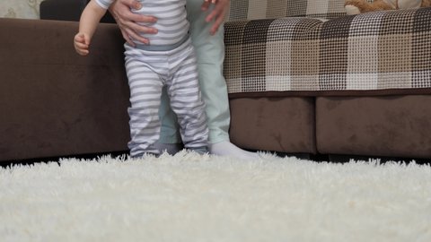 Happy mom holding hand helps cute little baby take first steps at home. The concept of childhood and a happy family. Infant barefoot girl boy learns to walk while standing on a warm floor.