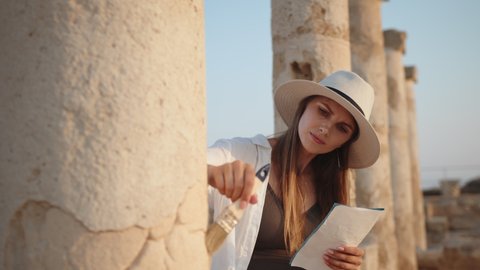 Archeologist standing among ruins of ancient temple. Woman holding brush for digging and map in hands. Girl In an ancient temple with columns looking at records of a map. Archaeological Digging Site.