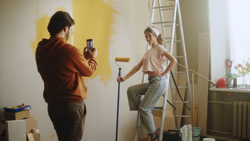 Young couple husband taking photos of wife on smartphone while making overhaul. Man and woman doing renovating together, painting walls in yellow. Interior design, new apartment.  Royalty-Free Stock Footage #1084228576