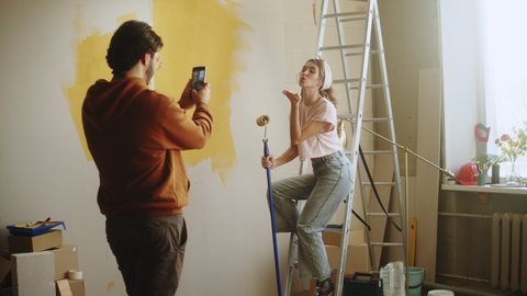 Young couple husband taking photos of wife on smartphone while making overhaul. Man and woman doing renovating together, painting walls in yellow. Interior design, new apartment. 