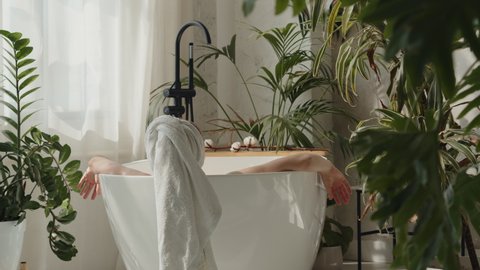 A young woman with towel on her head takes a bath in a luxury eco spa with lots of tropical plants.