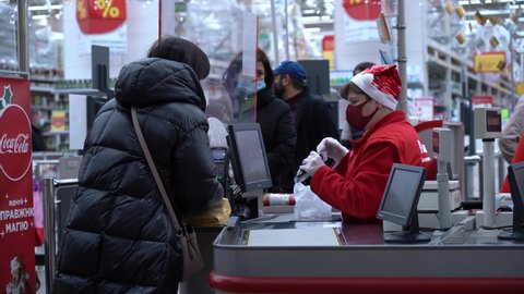 Kyiv, Ukraine - December 21, 2021: The buyer pays for purchases at the checkout in the supermarket on the eve of the New Year holidays. Cashier in a protective mask and Santa hat.