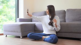 Glad Asian woman with a pleasant smile, waves with a hand at camera of cell phone in home interior, makes video call to her best friend sitting on a floor 