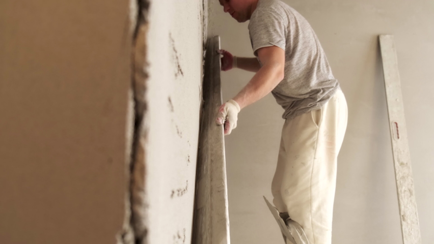 Male Builder Levels Plaster On Wall. Smooth Board. Removes Extra Layer. Renovation work in Residential Premises. Neat Movement. Profession. Thick Layer of Plaster. Preparing Walls for Whitewashing