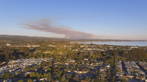 Aerial shot flying over suburbs at sunrise in Watsonville. Streets glistening from rays falling over trees and houses with a lake in the background. Smoke coming out of forest fire in the background.