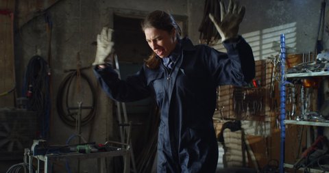 Cinematic shot of young happy smiling female mechanic in uniform is putting on smartphone her favorite music and dancing carefree while having break from working in maintenance repair workshop.