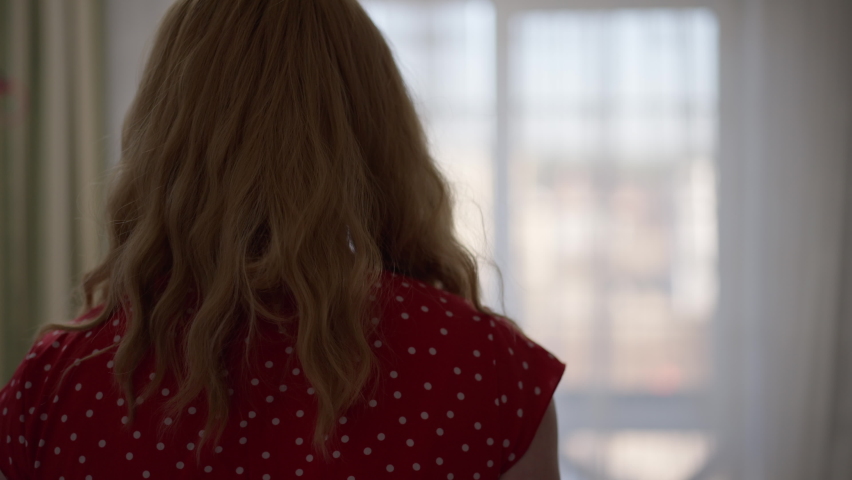 Back view confident trans woman walking to window in bedroom looking out and turning looking at camera smiling. Caucasian LGBT queer person in dotted red dress posing at home indoors