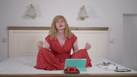 Wide shot front view relaxed trans woman meditating with eyes closed sitting on bed at home. Portrait of confident Caucasian LGBTQ non-binary person in lotus pose indoors in bedroom - Powered by Shutterstock - Get 15% off with code: PIKWIZARD15