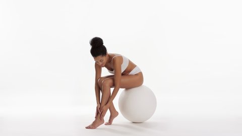 African American young slim attractive dark-haired woman in white underwear strokes her leg and puts her hand on her waist sitting on big white ball on white background | Wax commercial