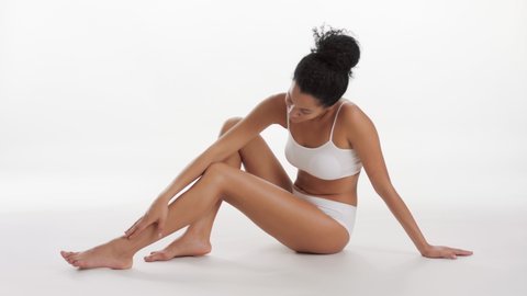 Horizontal long shot of African American slim gorgeous brunette woman in white underwear strokes her leg and looks at the camera smiling on white background | After leg shaving concept