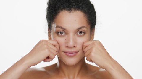 African American young slim good-looking black-haired woman with nude shoulders stretches her cheeks and smiles wide for the camera on white background | Skin stretching concept