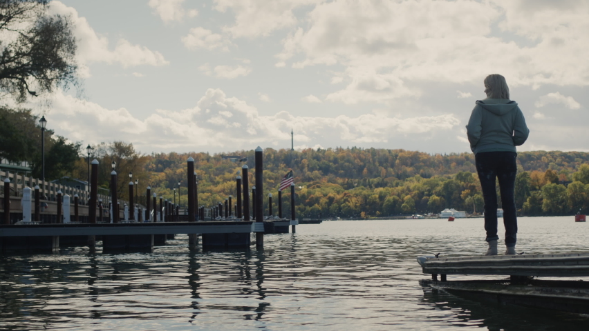 A woman stands on a pier overlooking Lake Ontario, the American flag is visible in the distance, there are no other people and yachts around. Autumn and end of tourist season.