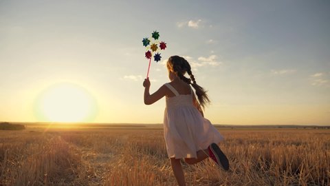 Happy girl runs across field at sunset with windmill in her hands. Silhouette of joyful girl holding wind toy. child enjoys fresh air. fun game in wind in field. Happy child runs at sunset.Wind toy