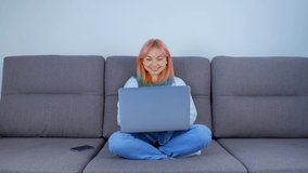 Emotional girl with dyed hair filming video call on laptop web camera. Cute young woman in late 20s calling her friends on internet with notebook computer while sitting on couch at home on lockdown