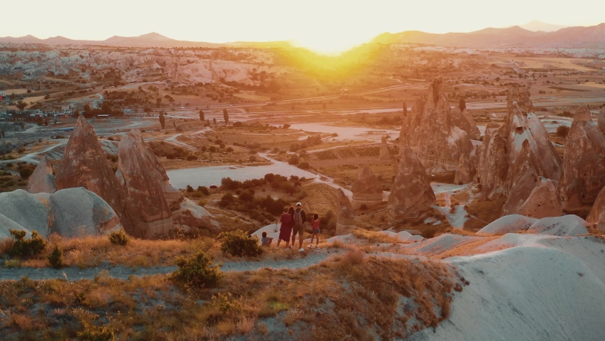 Drone flies around happy family with kids watching epic sunrise scenery panorama on top of mountain rock in Cappadocia.