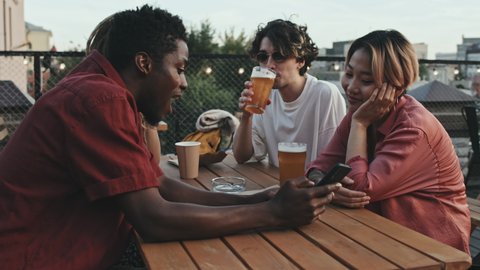 Medium shot of four multiethnic hipster friends drinking beer and chatting sitting at outdoor rooftop cafe on warm summer evening